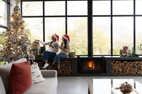 Diverse women wearing santa hats sipping hot drinks by fireplace beside christmas tree