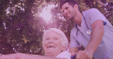Senior woman smiling while caregiver pushing wheelchair under sunlit tree canopy