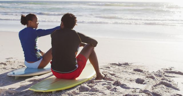 A couple in swimwear sitting on surfboards while relaxing on the beach, watching the ocean waves. Suitable for themes on summer activities, romance by the sea, surfing experiences, and beach relaxation.