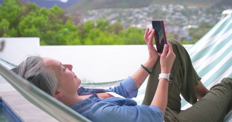 Senior woman relaxing in hammock on sunny balcony, using smartphone and smartwatch