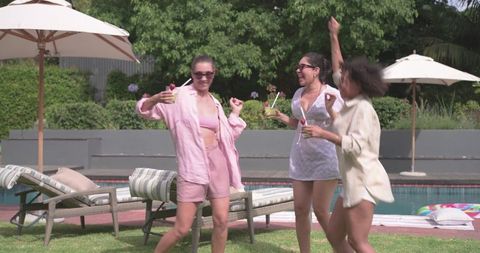 Diverse women celebrating by pool clinking cocktails and laughing at summer backyard party