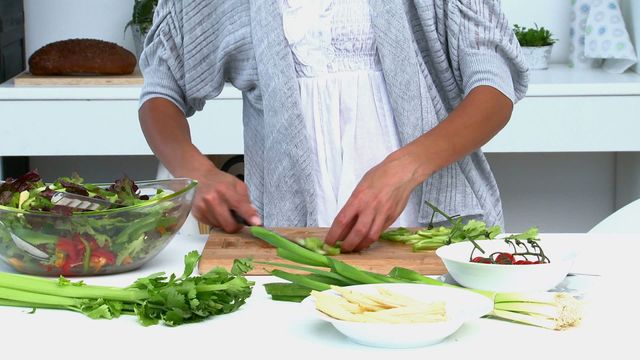 A woman is preparing a variety of fresh vegetables on a cutting board in a modern kitchen environment. This captures a focus on healthy eating as she expertly slices produce like greens and peppers, with a ready-to-toss salad in clear view. Ideal for use in content promoting nutrition, wellness courses, healthy lifestyle blogs, or cooking classes.