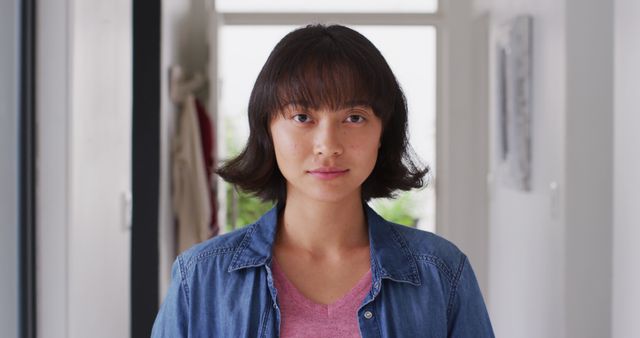 Young woman stands confidently indoors wearing denim shirt and pink top. She has short hair with bangs and looks directly at the camera. Suitable for themes related to youth, confidence, casual fashion, home interior, and personal portraits.