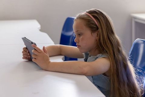 Schoolgirl focusing on tablet while leaning on white desk in classroom with blue chairs