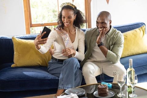 Couple celebrating birthday at home taking selfie on sofa with cake and wine