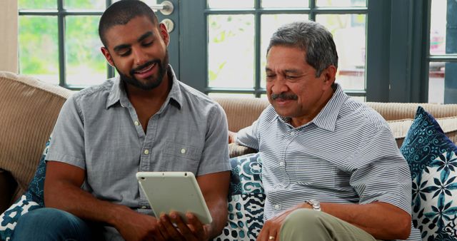 Younger man teaching older man to use a tablet while sitting on a couch in a well-lit living room. Both are smiling, creating a friendly and cooperative atmosphere. Useful for depicting technology education, digital assistance, multigenerational interaction, and family bonding in technological contexts.