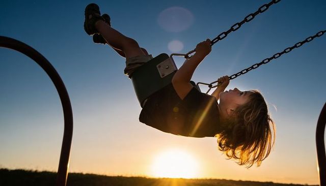 Backlit child swinging at sunset on green bucket swing, golden hour playground joy, playful motion