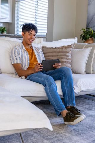 Adult asian man relaxing on white sofa smiling while using tablet in bright living room