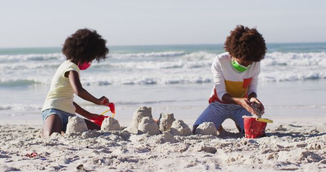 Two children wearing face masks building sandcastles on a beach near the ocean, practicing teamwork and creativity. Use for scenes depicting summer fun, outdoor family activities, or health-conscious social interactions during pandemic.