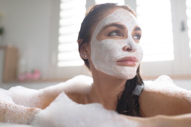 Woman enjoying a relaxing bath with a white face mask on her face. Great for topics on self-care, skincare routines, beauty treatments, and spa days. Use it in articles, blog posts, and promotional materials for beauty and wellness products.