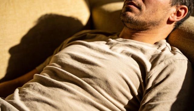 Reclining man wearing light t-shirt on sunlit beige couch showing fabric folds and shadowed silhouet