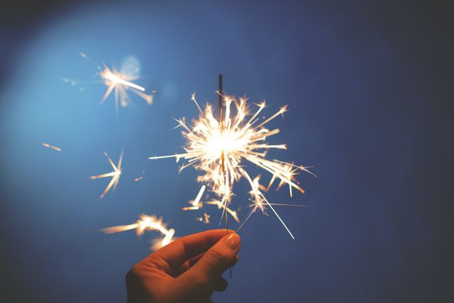 Hand holding lit sparkler in foreground with blue background. Perfect for themes of celebration, festive events, parties, New Year's Eve, Fourth of July, and other joyous occasions. Can also be used to illustrate concepts of light, fun, and excitement.