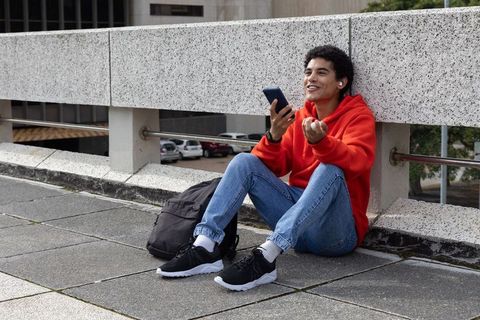 Young man sitting on urban walkway using smartphone and earbuds wearing red hoodie