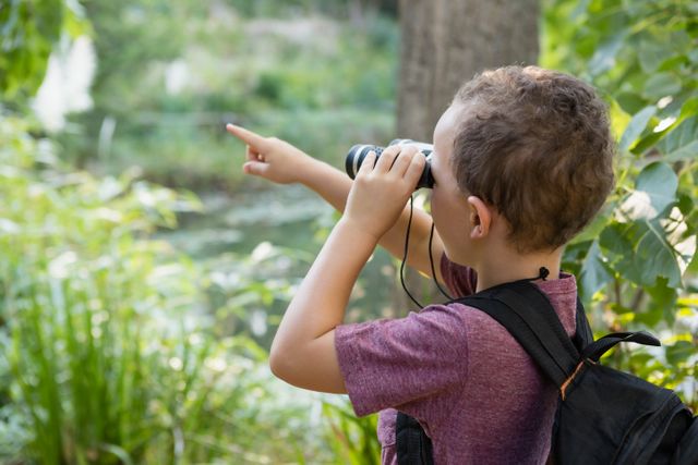 Young boy with binoculars and backpack pointing at something in the forest. Ideal for educational content, nature exploration themes, children's activities, and outdoor adventure promotions.