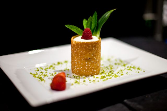 Elegant gourmet dessert served on white plate. Delicate pastry cylinder filled with cream, topped with fresh raspberry and mint leaves, with crushed nuts and strawberry slice garnish. Ideal for culinary blogs, restaurant menus, food-related content, and fine dining promotions.