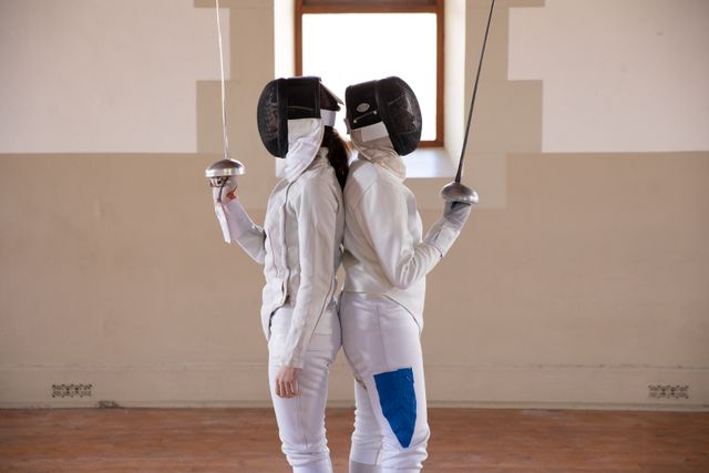 Two people preparing for a fencing duel, wearing protective fencing clothes, holding sabers, in a gym. Sport and working out.
