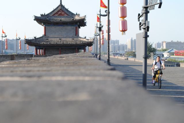 Man cycling on the ancient city wall in Xi'an, adorned with red lanterns and traditional Chinese architecture. Urban skyline visible in the background. Ideal for use in travel brochures, cultural heritage articles, tourism marketing, and advertisements for historical sites.