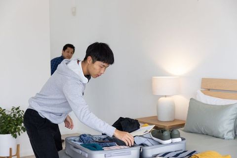 Asian teen packing suitcase on bed while father watching, preparing for family trip