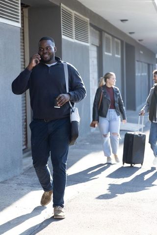 Urban commuters walking with luggage and coffee, man talking on phone on sunlit sidewalk