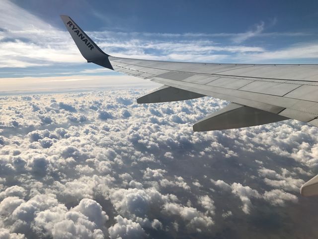 View of airplane wing from passenger window with blue sky and clouds below. Perfect for travel-related articles, aviation promotions, and content about flight experiences. Could be used in blogs, promotional materials, and advertisements.