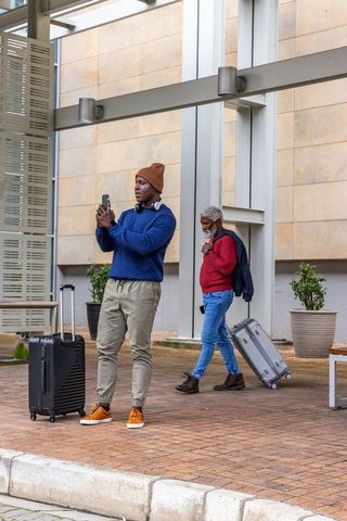 African american man texting while waiting with luggage, older traveler pulling suitcase