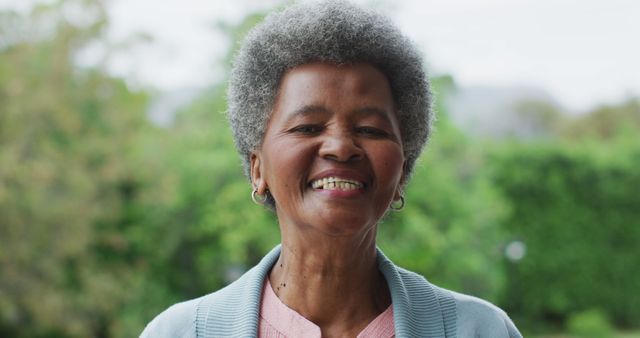 Elderly woman with grey hair, smiling warmly while standing outdoors against lush, green background. Ideal for use in retirement, health, wellness, or senior living advertisements.
