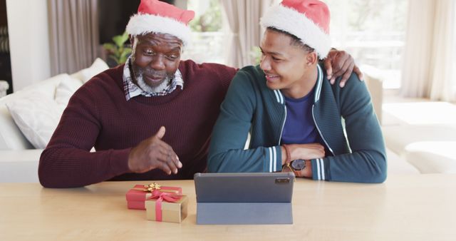 African American father and son wearing Santa hats sitting at table with tablet and Christmas gifts. Ideal for holiday season advertisements, family bonding themes, and promoting festive spirit. Perfect for technology-related holiday promotions and father-son relationship topics.