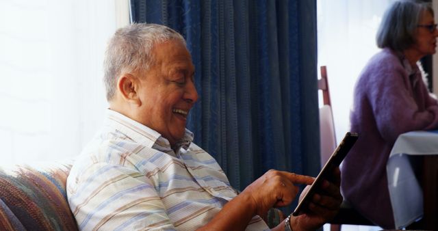 Elderly man is sitting on a couch using a tablet and smiling happily. Well-lit living room with cozy ambiance. Could be used in articles or advertisements about senior technology adoption, retirement contentment, or home interior comfort for seniors.