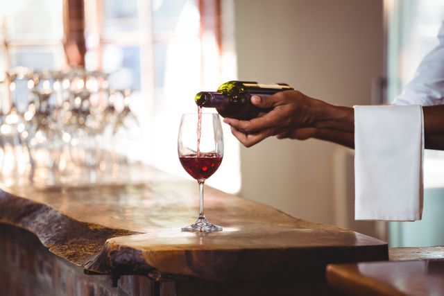 Bartender pouring red wine into a glass at a bar counter. Ideal for use in hospitality industry promotions, restaurant and bar advertisements, wine tasting events, and luxury dining experiences.
