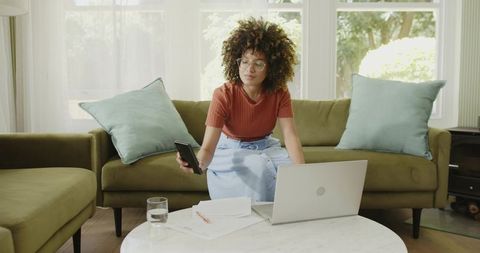 African American woman working remotely on laptop checking phone in bright living room