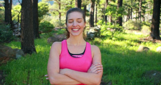 Woman in bright pink tank top stands with arms crossed while smiling, surrounded by lush forest greenery and illuminated by sun. Ideal for health and wellness content, outdoor activities promotion, fitness advertisements, or positive mental health campaigns.