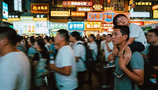 Asian father carrying sleeping child on shoulders walking through busy neon night market