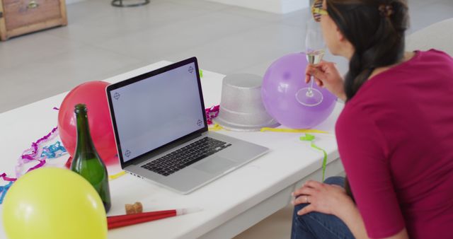 Woman enjoying a virtual party, celebrating with friends online via video call. She is holding a glass of champagne and there are colorful balloons, party hats, and streamers on the table. Suitable for illustrating remote celebrations, online social gatherings, virtual events, and digital engagements.