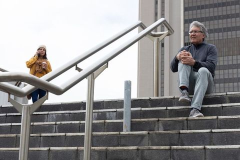 Senior man sitting on concrete steps holding knee near metal handrail looking thoughtful