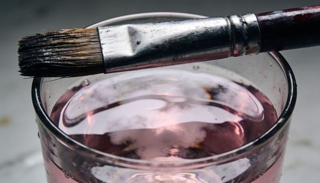 Flat paintbrush resting on glass rim with pink rinse water, macro studio still life
