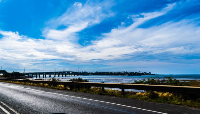 This image captures a scenic view from a highway overlooking the ocean with a prominent bridge in the distance under a vibrant blue sky. Perfect for travel websites, transportation blogs, coastal real estate promotions, and inspirational posters aimed at adventure and road trips.