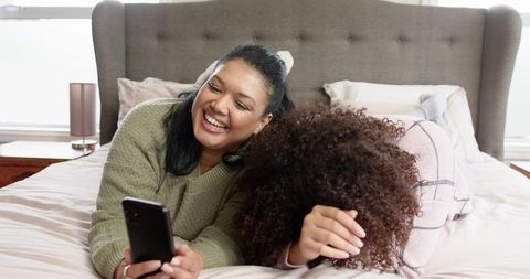 Mother daughter laughing on bed holding smartphone cozy family bonding candid moment