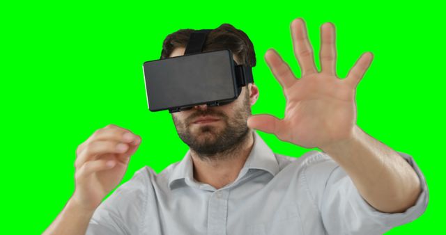 Man wearing VR headset appears deeply engaged with virtual environment, hand lifted as if interacting with unseen elements. Green screen background allows easy adjustment for diverse uses. Ideal for technology promotions, virtual reality advertisements, digital simulations, and gaming industry demonstrations.