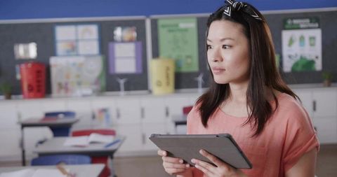 Female teacher holding tablet and surveying empty elementary classroom for lesson planning