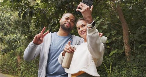 Couple taking selfie on park trail in activewear while holding yoga mat and water bottle