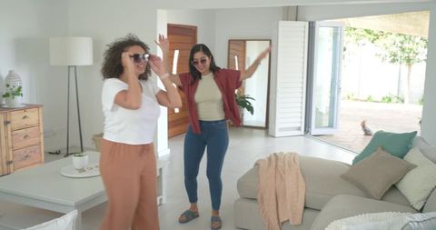 Diverse female friends dancing and laughing in bright modern living room with sunglasses