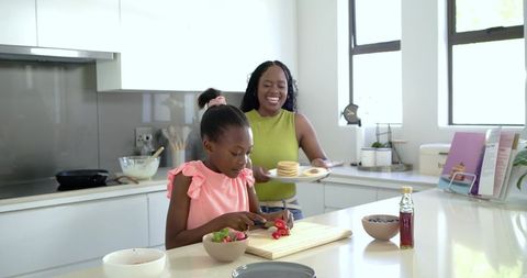 African american mother serving pancakes while daughter chopping strawberries in sunlit kitchen
