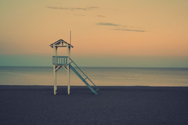 A quiet sunrise is casting soft pastel colors over an empty lifeguard tower on a serene beach. This can be used for concepts related to peaceful beginnings, solitude, and the beauty of nature at dawn.