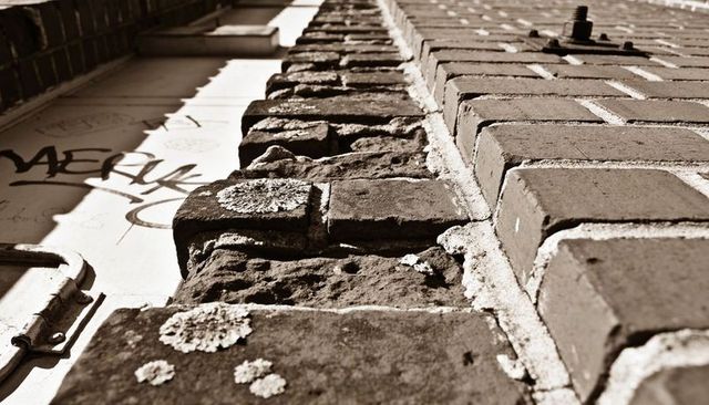 Low-angle weathered brick and stone wall with lichen, graffiti, rusted bolt, sepia
