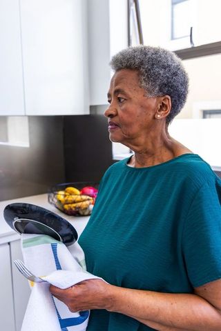 Senior african american woman drying dishes in modern kitchen holding fork with fruit bowl