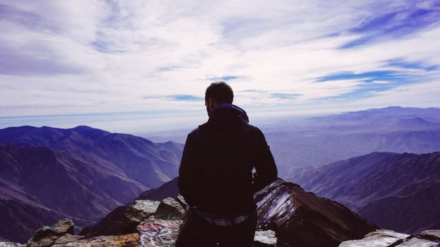 A man stands on a mountain peak with his back to the camera, looking out over a wide expanse of rugged mountain ranges. The sky is partially cloudy, adding texture to the vast landscape below. Perfect for depicting themes of adventure, exploration, and the transformative power of nature. Ideal for travel blogs, adventure magazines, social media content relating to hiking or mountaineering, and advertisements promoting outdoor gear or experiences.