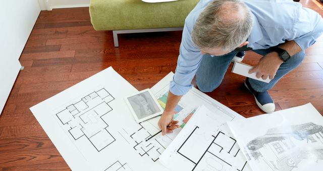 An architect is reviewing house plans and blueprints spread on a wooden floor. Ideal for articles or websites discussing architecture, house construction, interior design projects, or professional planning processes. Useful for illustrating topics relating to creative workspace, engineering, and project management.