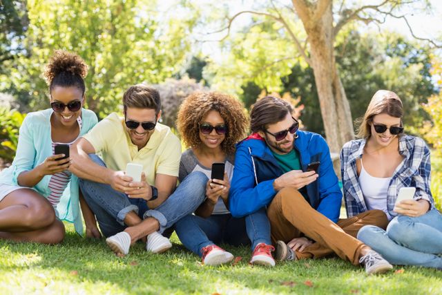 Group of friends sitting on grass in park, using smartphones. Ideal for themes related to social media, technology, outdoor activities, friendship, and leisure. Perfect for advertisements, blog posts, and articles about modern communication, youth culture, and outdoor fun.