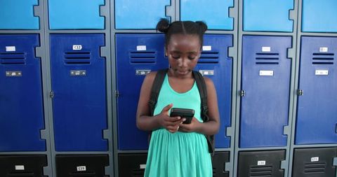 African schoolgirl standing at blue lockers using smartphone in teal dress