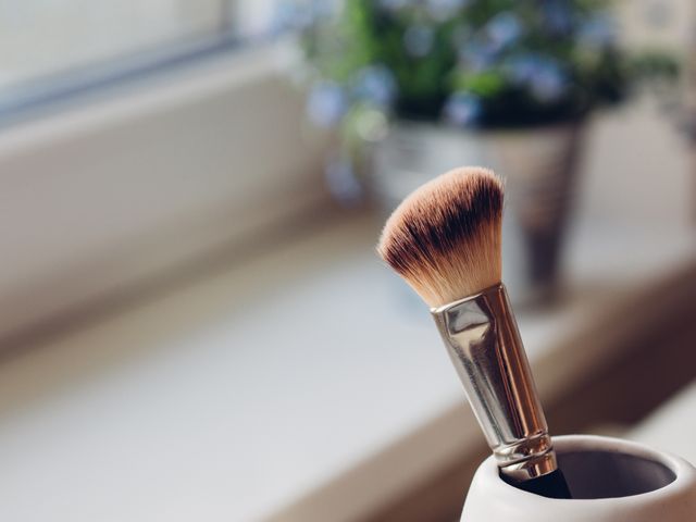 This detailed close-up of a makeup brush with softly blurred bristles perfectly captures a tranquil beauty routine. The placement on a windowsill with a slight glimpse of a potted plant in the background illuminated by natural light suggests a serene morning. Ideal for use in blogs, articles, or advertising focused on beauty, self-care, home decor, and lifestyle.